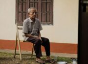 Elderly man sitting pensively on a wooden chair outside his home, reflecting solitude.